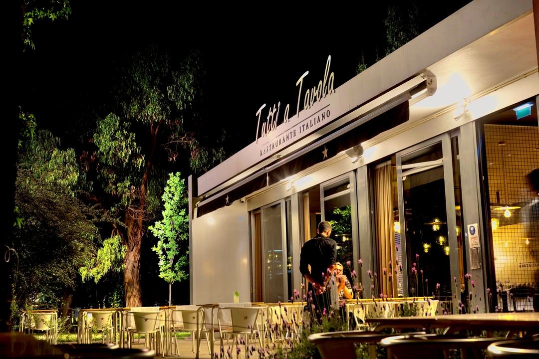 A fachada iluminada do Tutti a Tavola, à noite, entre as árvores